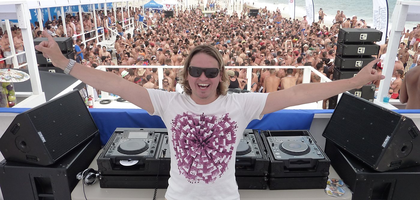 Lee Dagger, DJ Bimbo Jones, arms out behind the decks in front of a packed beach party